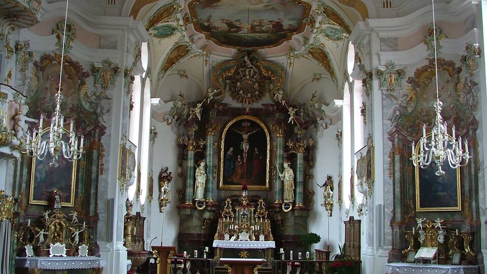An ornate church interior with elaborate ceiling frescoes, gilded altars, and chandeliers.