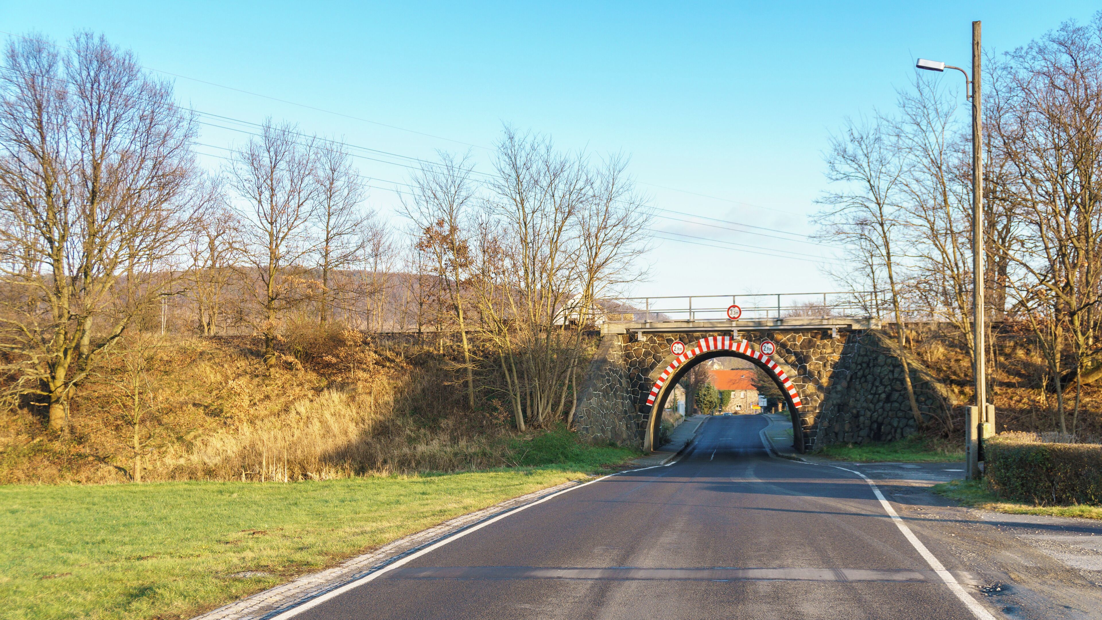 A rural road passing under a stone bridge with a height restriction sign.