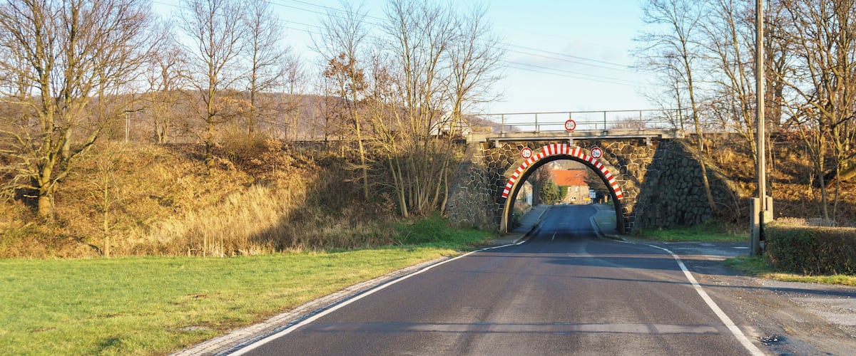 A rural road passing under a stone bridge with a height restriction sign.