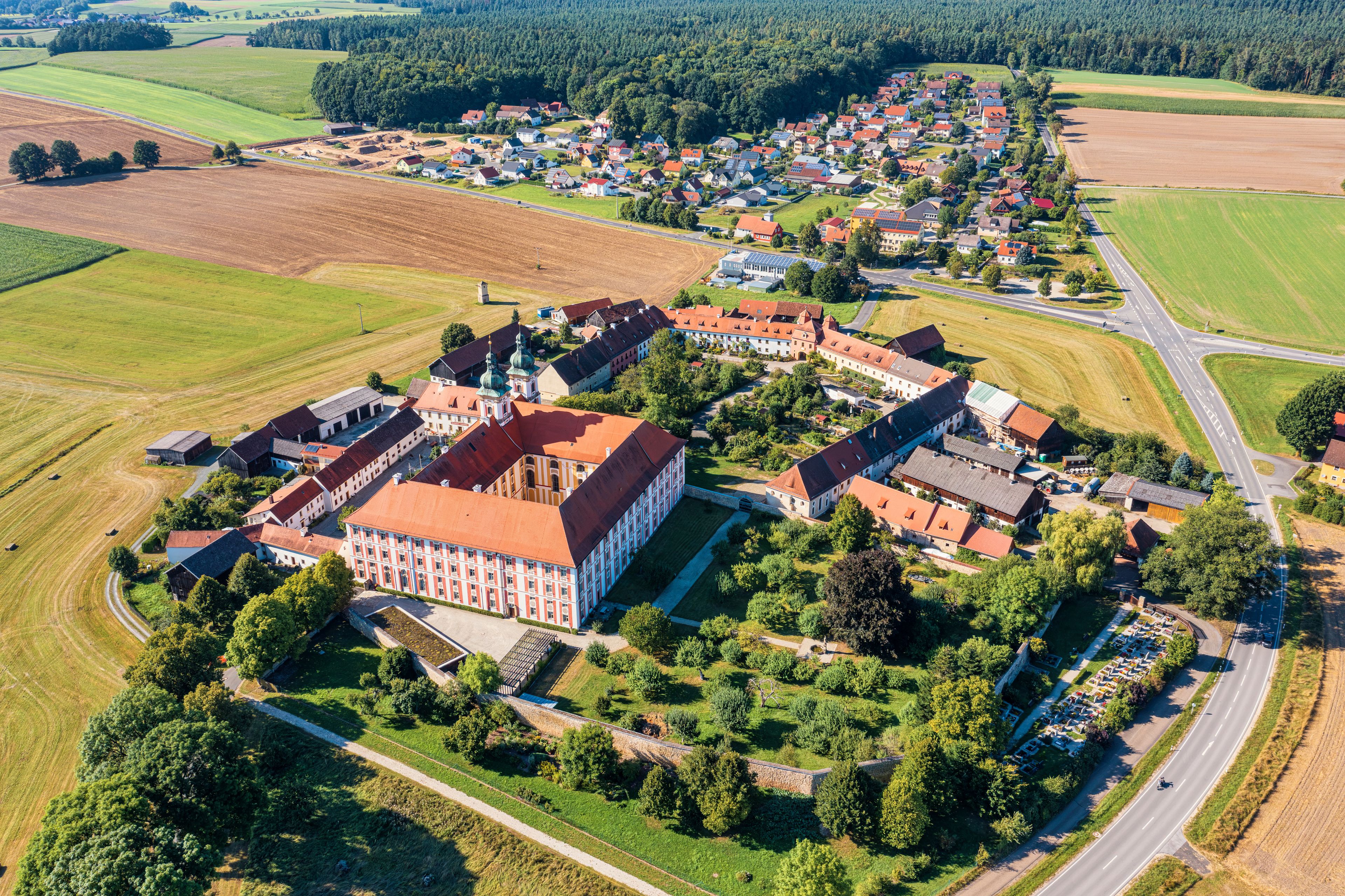 Vue aérienne d’une ville avec un bâtiment historique remarquable, entouré de champs et d’une forêt dense en arrière-plan.