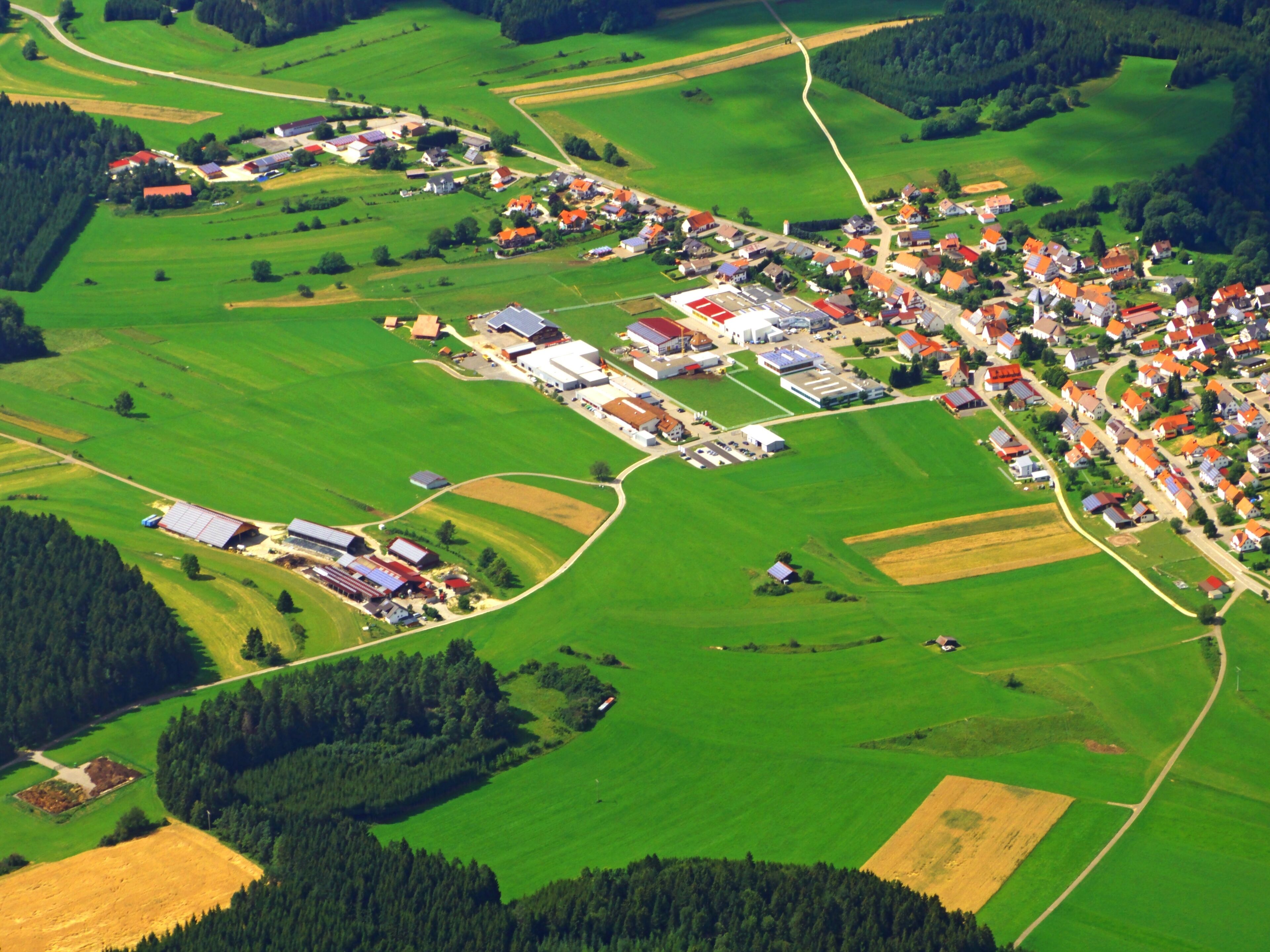 An aerial view of a rural village with green fields, a winding road, and a mix of residential and commercial buildings.