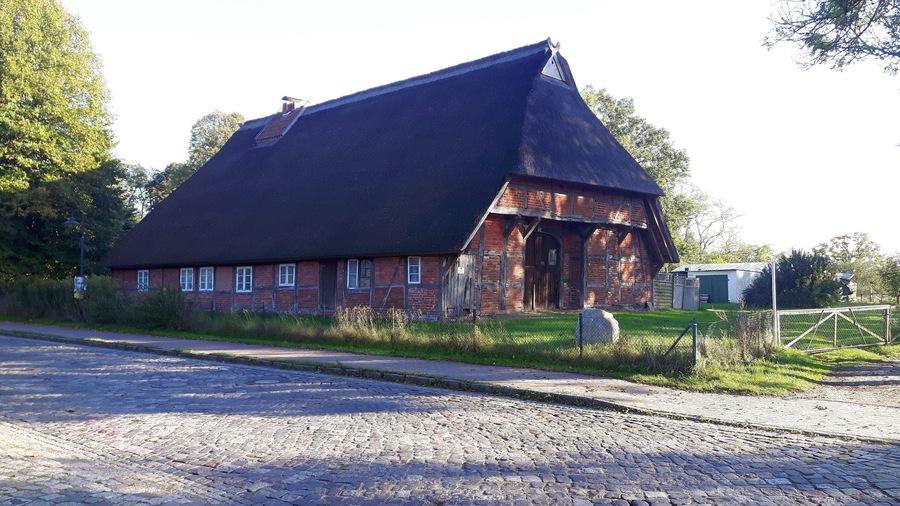 A traditional brick house with a steeply pitched roof, surrounded by a cobblestone road and greenery.