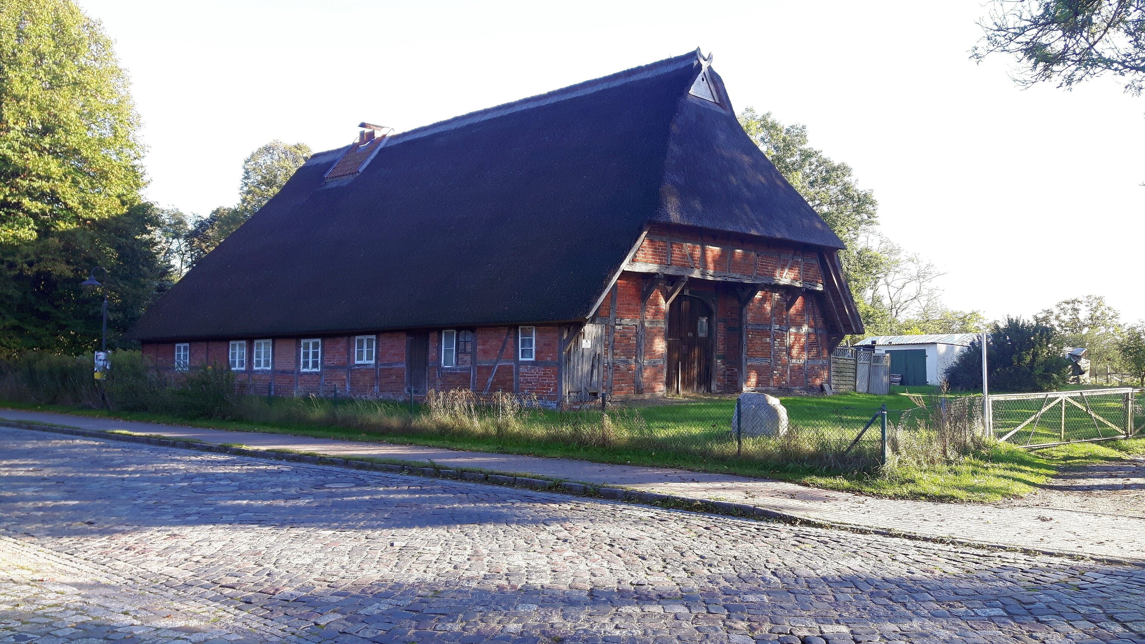 A traditional brick house with a steeply pitched roof, surrounded by a cobblestone road and greenery.