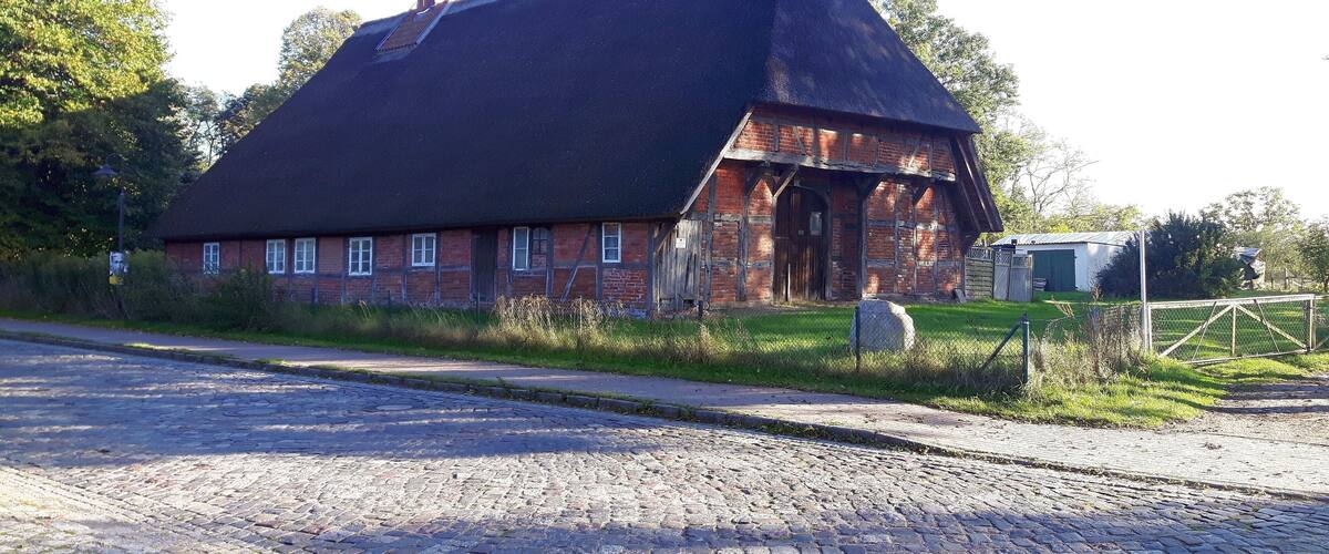 A traditional brick house with a steeply pitched roof, surrounded by a cobblestone road and greenery.