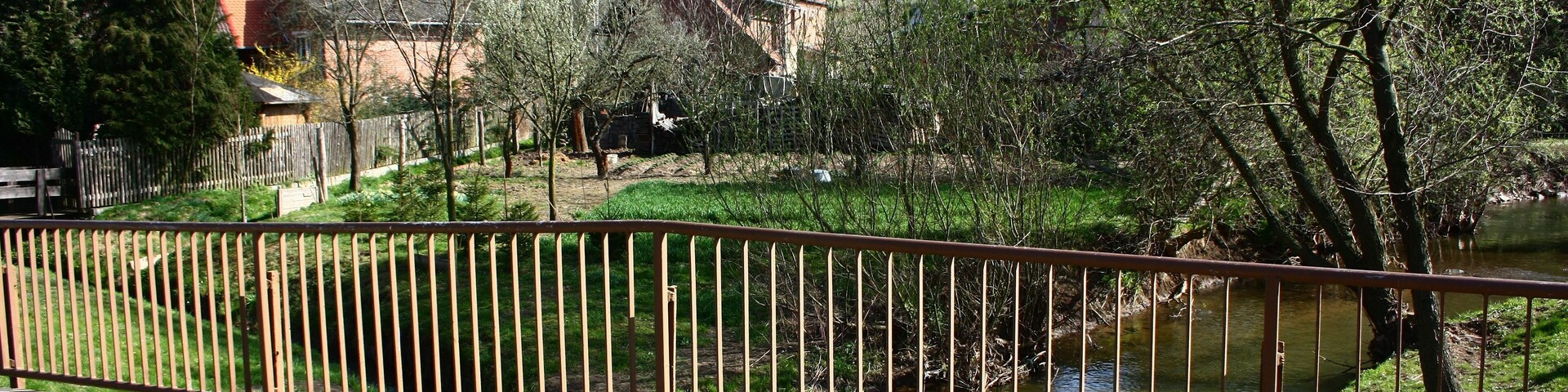 Un sendero pavimentado, una valla metálica, un pequeño arroyo y edificios al fondo.