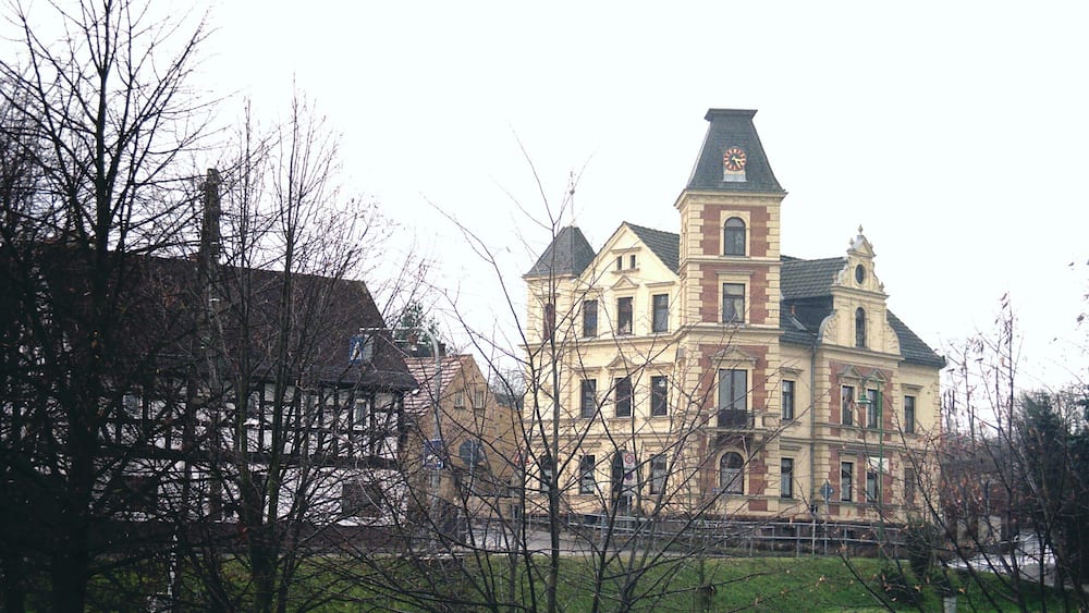 A large, historic building with a clock tower, surrounded by trees and a grassy area.