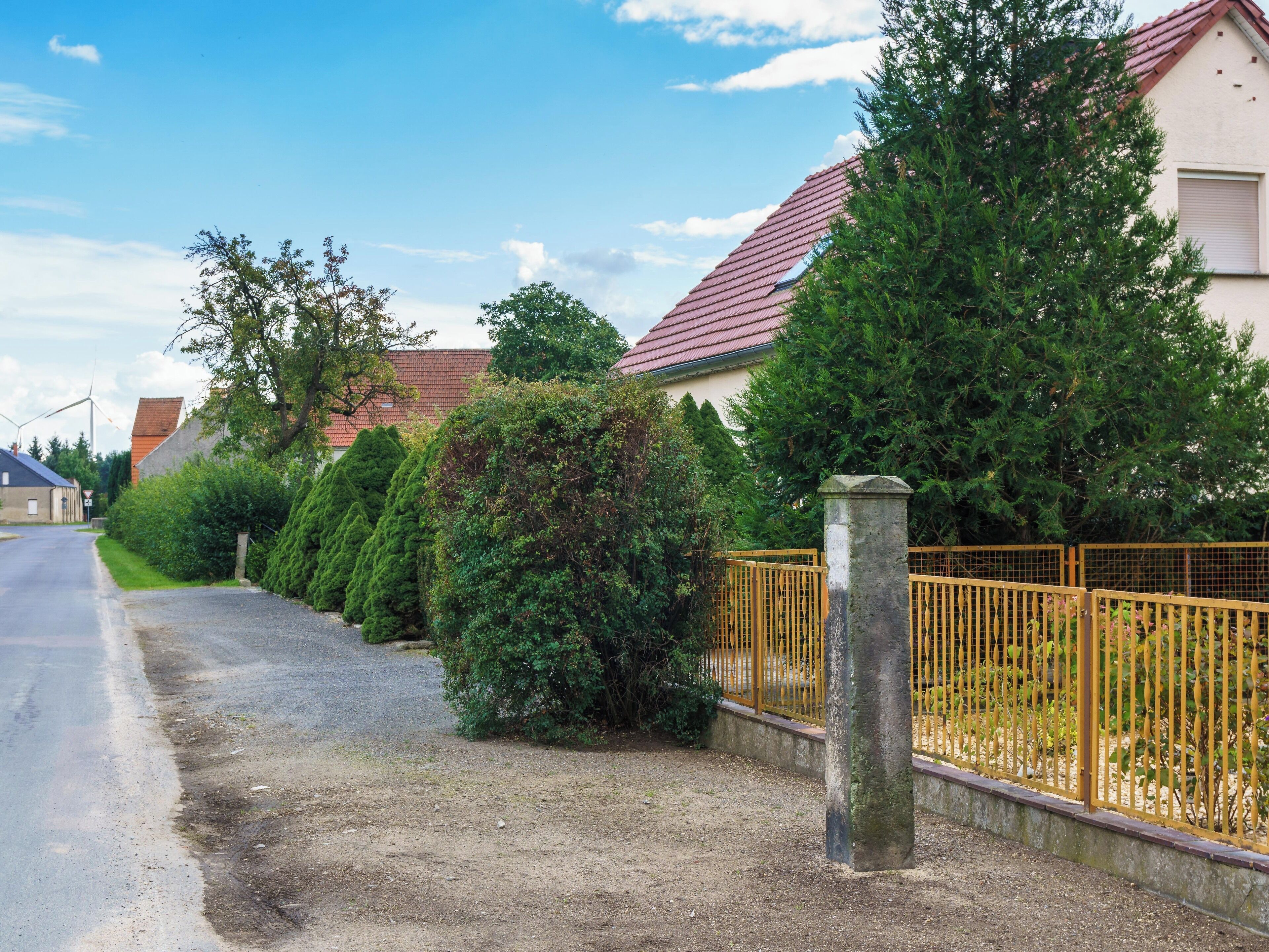 A residential street with a red-roofed house, a yellow fence, and a gravel path.