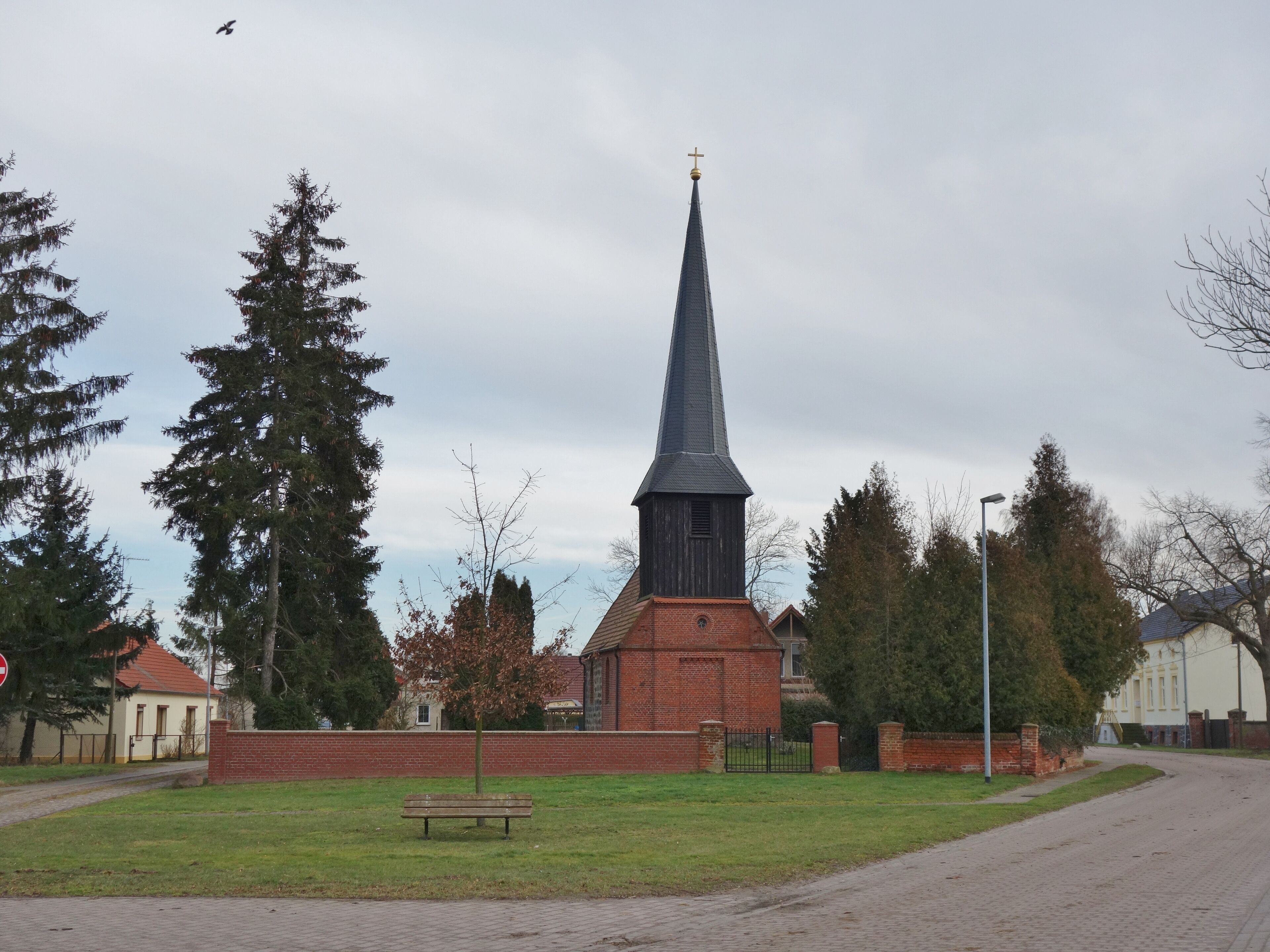 A church with a tall steeple and a cross on top, surrounded by trees and a bench in a grassy area.