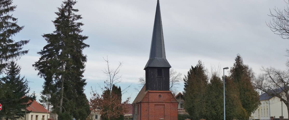 A church with a tall steeple and a cross on top, surrounded by trees and a bench in a grassy area.