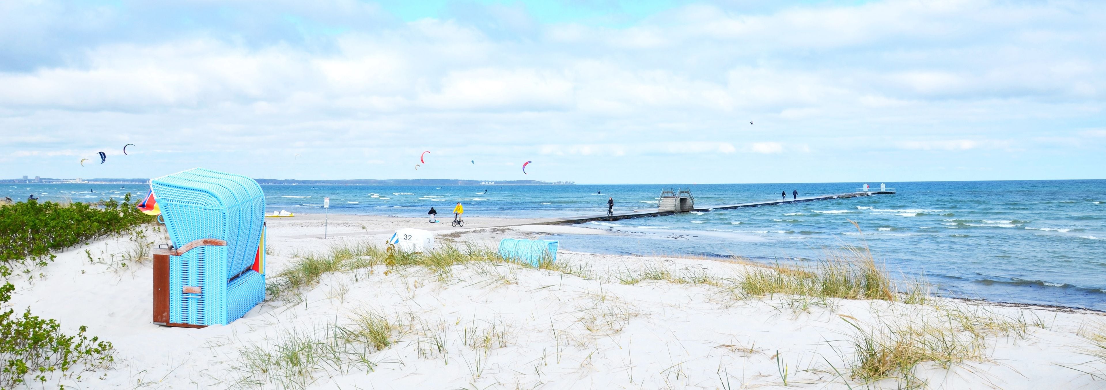 A beach with a jetty extending into the water, people walking and cycling, and kites flying in the sky.