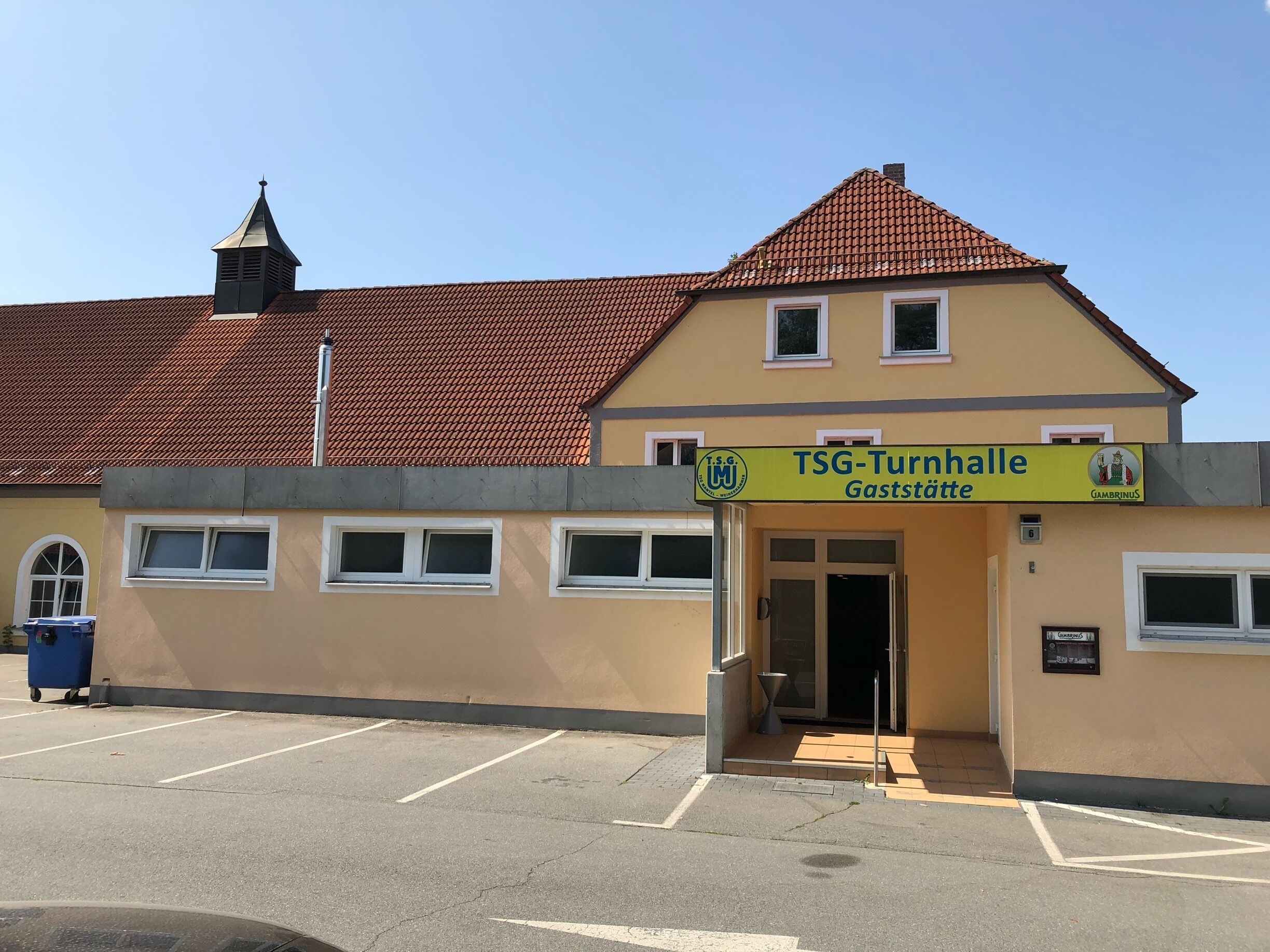 A building with a sign reading "TSG-Turnhalle Gaststätte" and a parking lot in front.