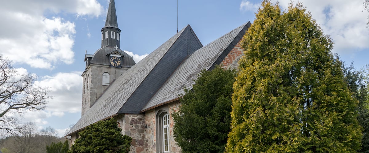A church with a steeple and a clock, surrounded by trees and a gravel path.