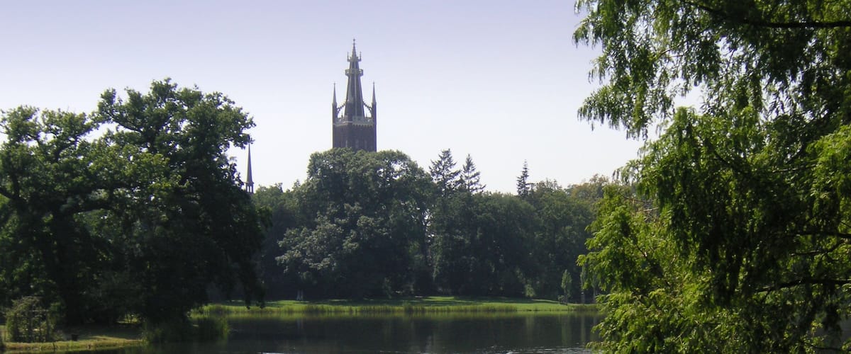 A calm lake with a boat carrying people, surrounded by lush greenery and a distant church tower.