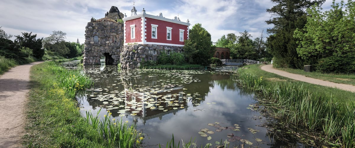 Un bâtiment historique doté d’une arche en pierre, d’un étang parsemé de nénuphars et d’un sentier.