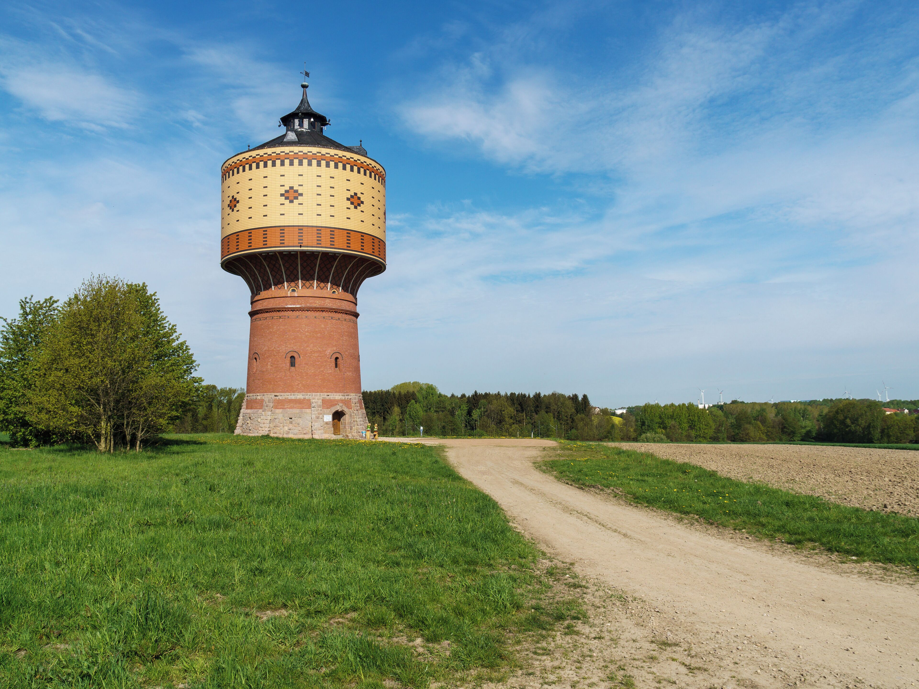 A historic water tower with a brick base and a tiled dome, surrounded by a grassy field and a dirt path.