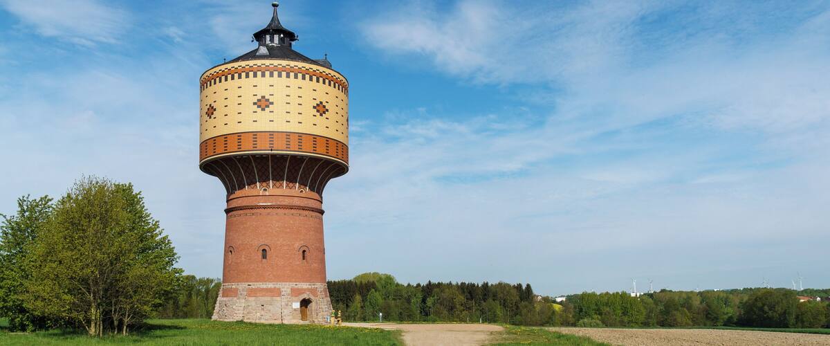 A historic water tower with a brick base and a tiled dome, surrounded by a grassy field and a dirt path.
