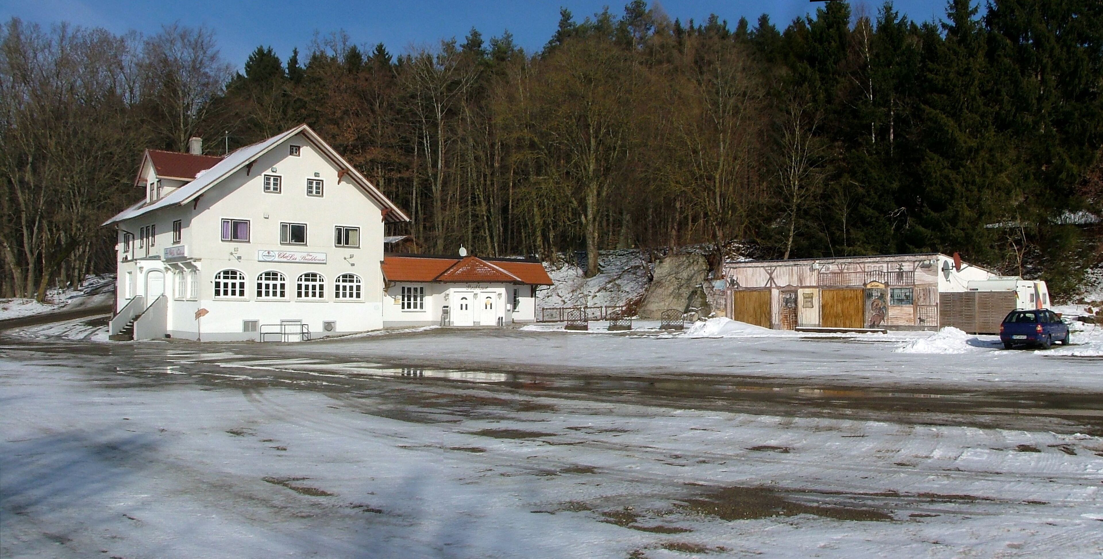 A white building with a red roof, a blue car, and a forest in the background.