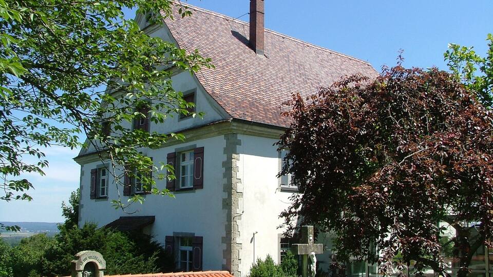 A white house with a red-tiled roof, surrounded by greenery and a clear sky.