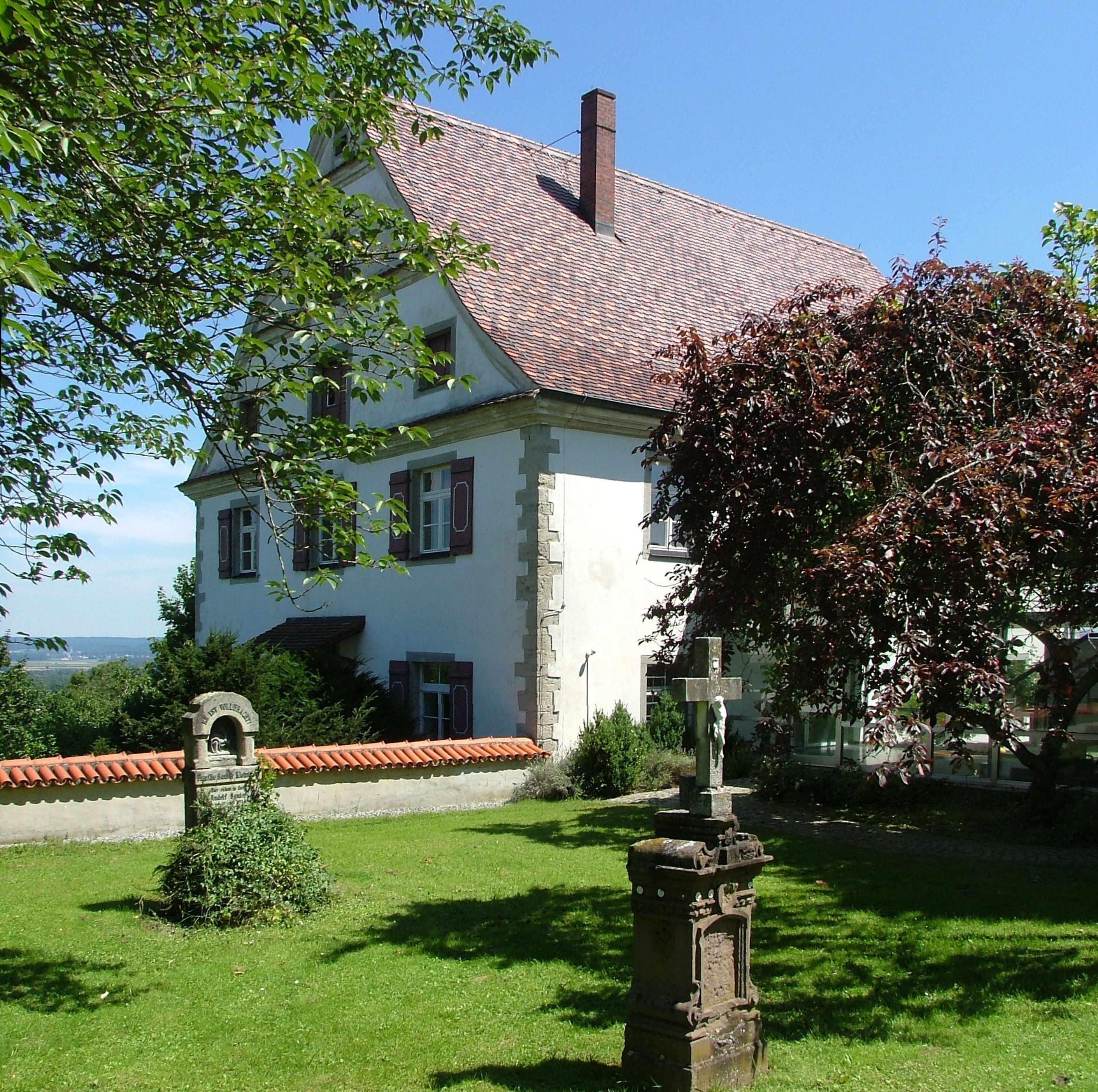A white house with a red-tiled roof, surrounded by greenery and a stone cross.