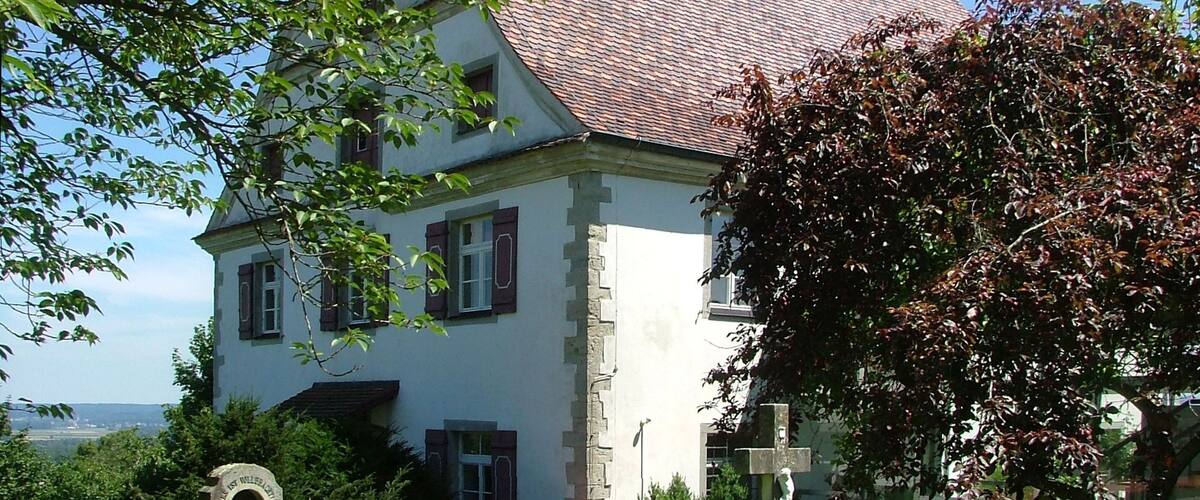 A white house with a red-tiled roof, surrounded by greenery and a stone cross.