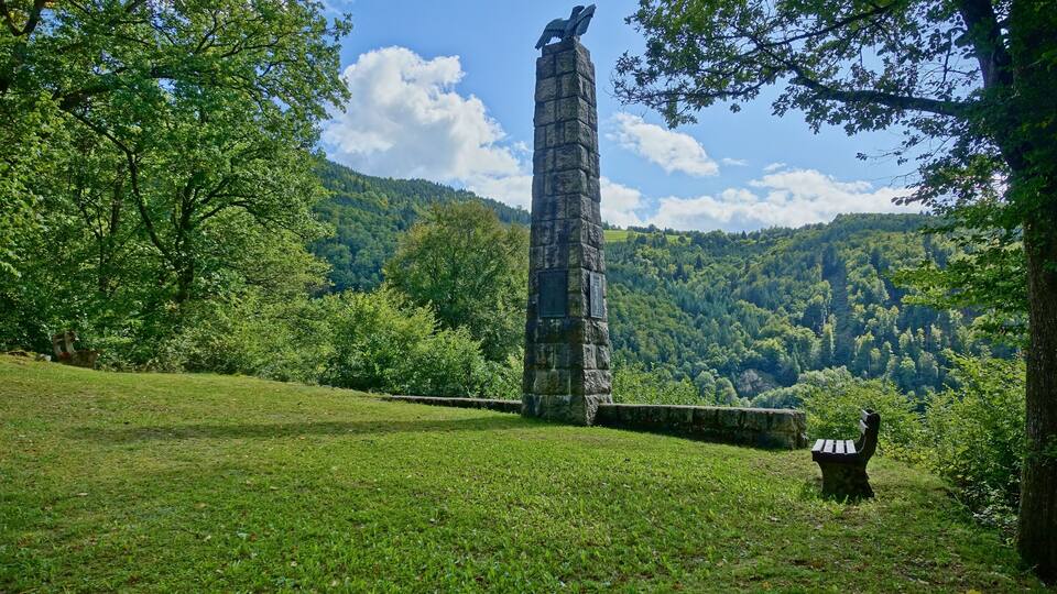 A stone monument with an eagle statue, surrounded by lush green trees and a mountainous landscape.