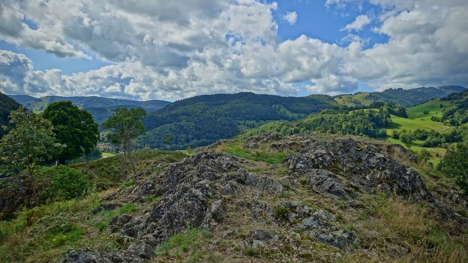 A mountainous landscape with rocky outcrops, dense forests, and rolling hills under a partly cloudy sky.