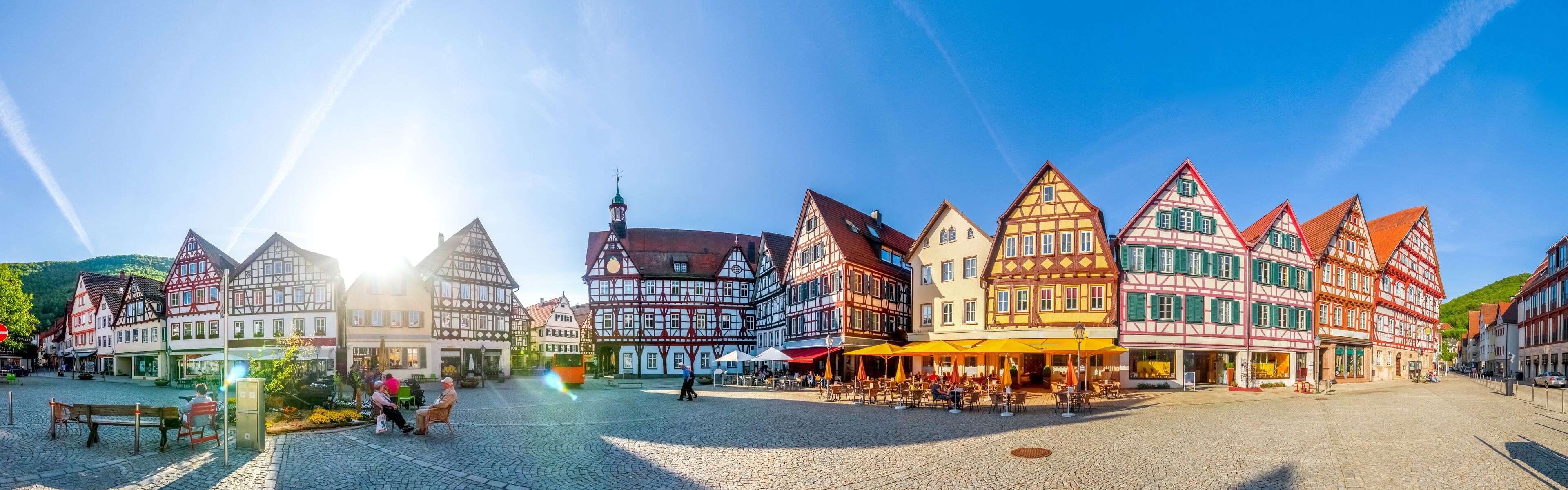 A cobblestone square with traditional half-timbered buildings, a tram, and outdoor seating areas.