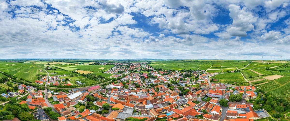 A panoramic view of a town with red-roofed buildings, surrounded by green fields and wind turbines in the distance.