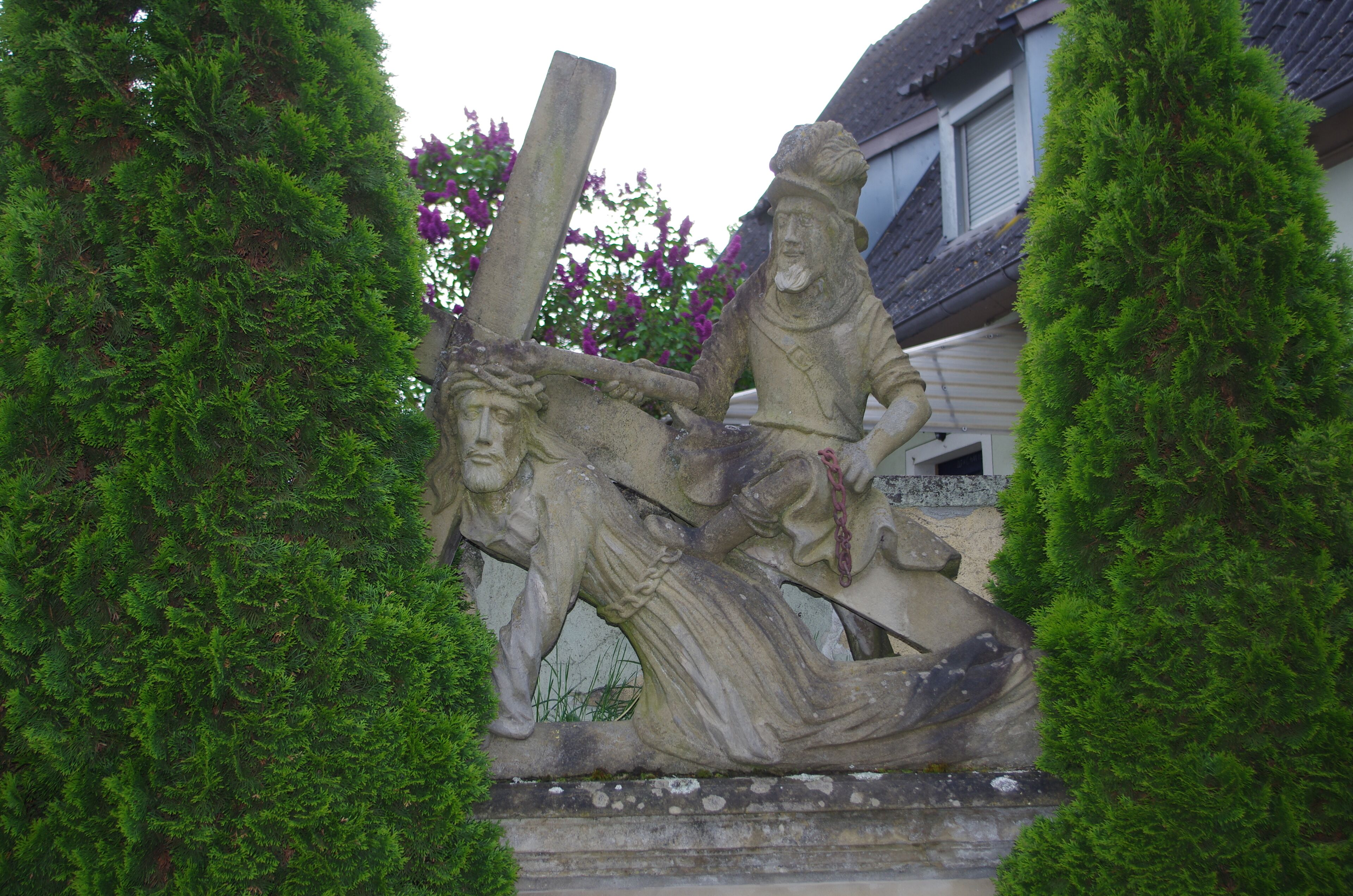A stone sculpture of a man holding a cross, surrounded by lush green trees.