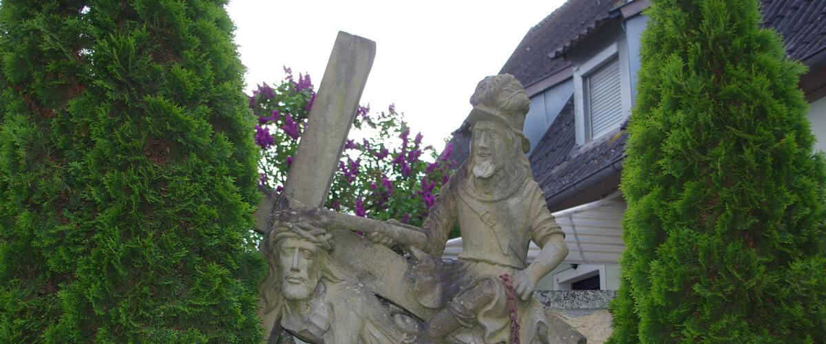 A stone sculpture of a man holding a cross, surrounded by lush green trees.