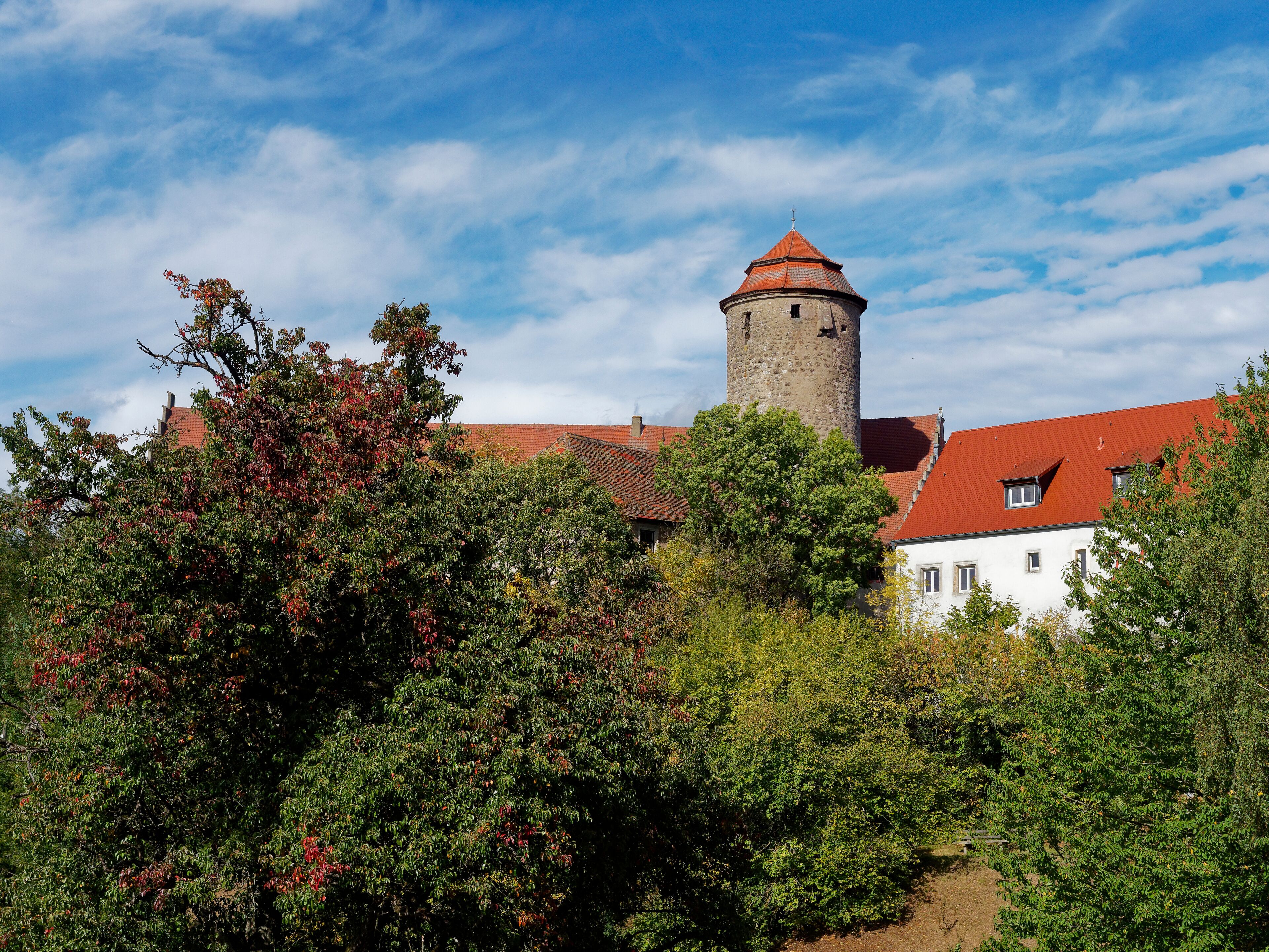 A historic tower with a red roof, surrounded by lush greenery and a white building with red-tiled roofs.