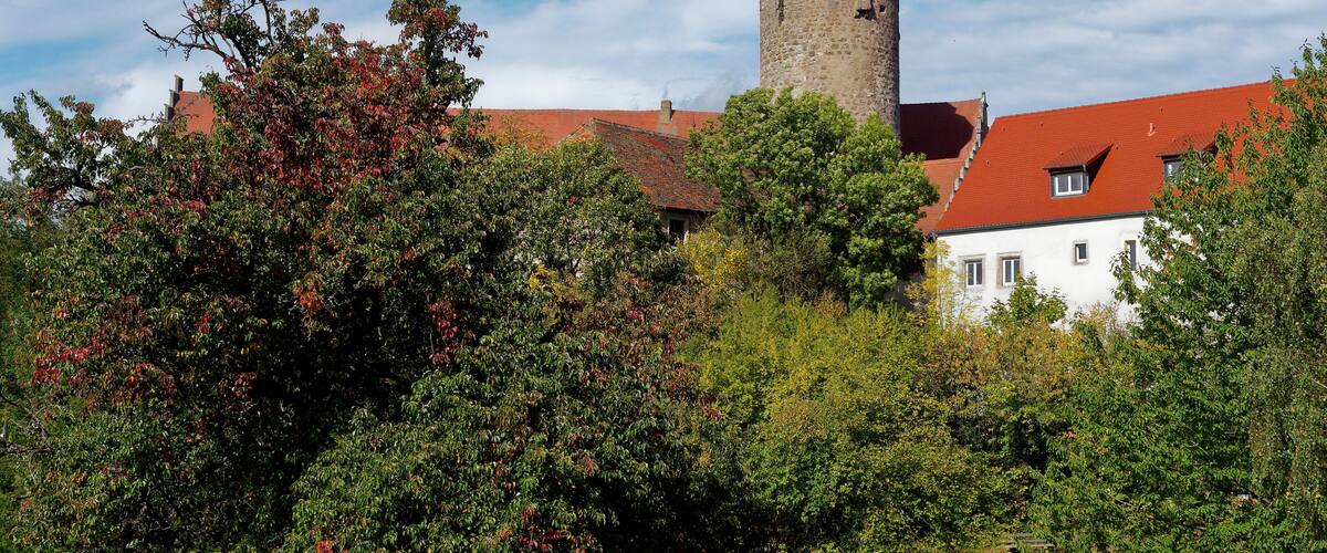 A historic tower with a red roof, surrounded by lush greenery and a white building with red-tiled roofs.