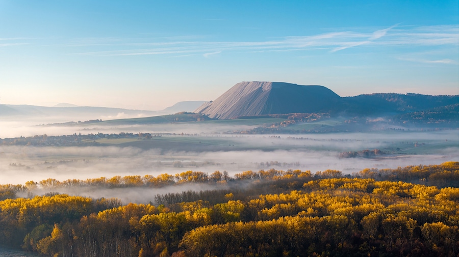 A mountain with a flat top, surrounded by autumn-colored trees and a layer of fog.