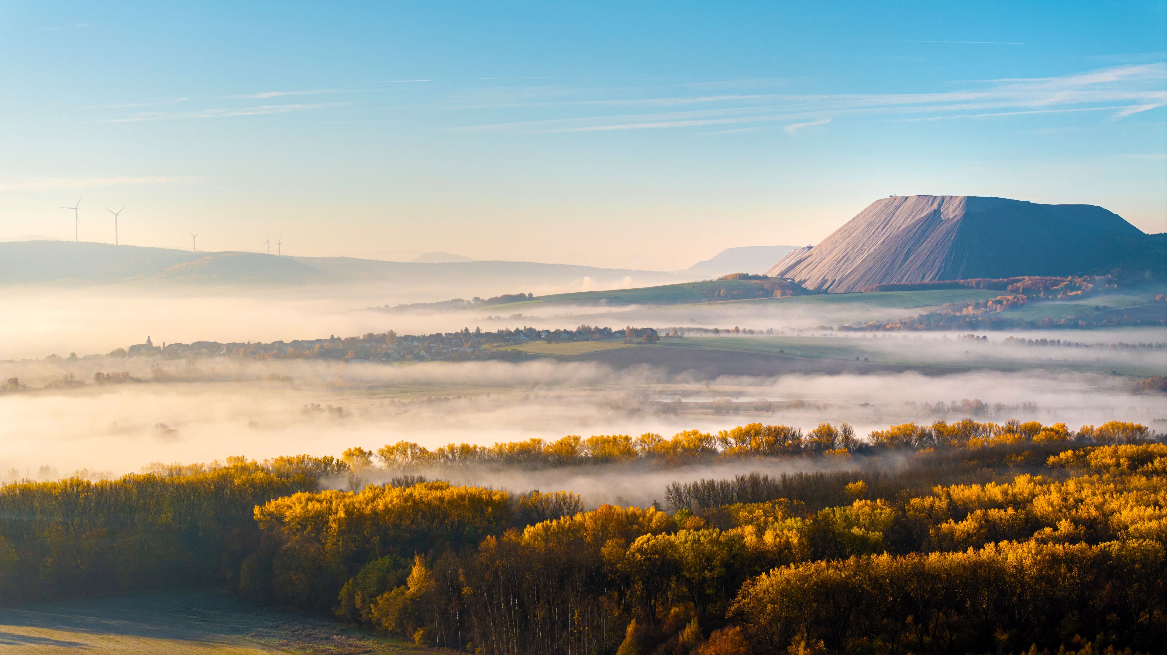Aerial view of the Monte Kali salt mountain rising above a layer of morning mist and golden autumn forests near the town of Heringen, Hessen, Germany.