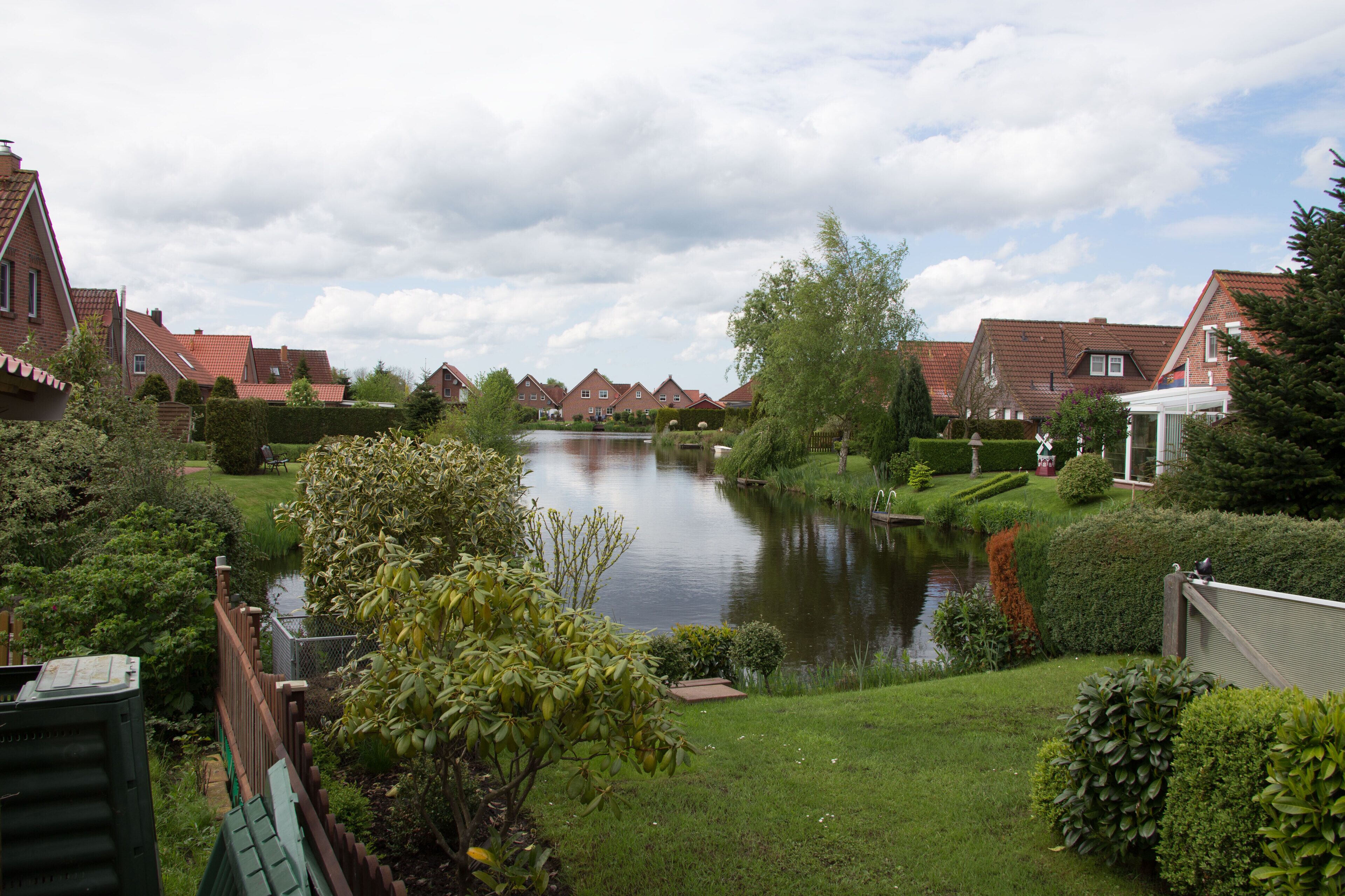 A serene riverside view with a row of red-roofed houses, lush greenery, and a calm water body.