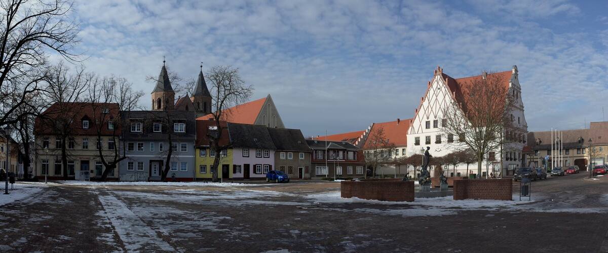 A snow-covered street with historic buildings, including a church with two spires and a triangular roof.