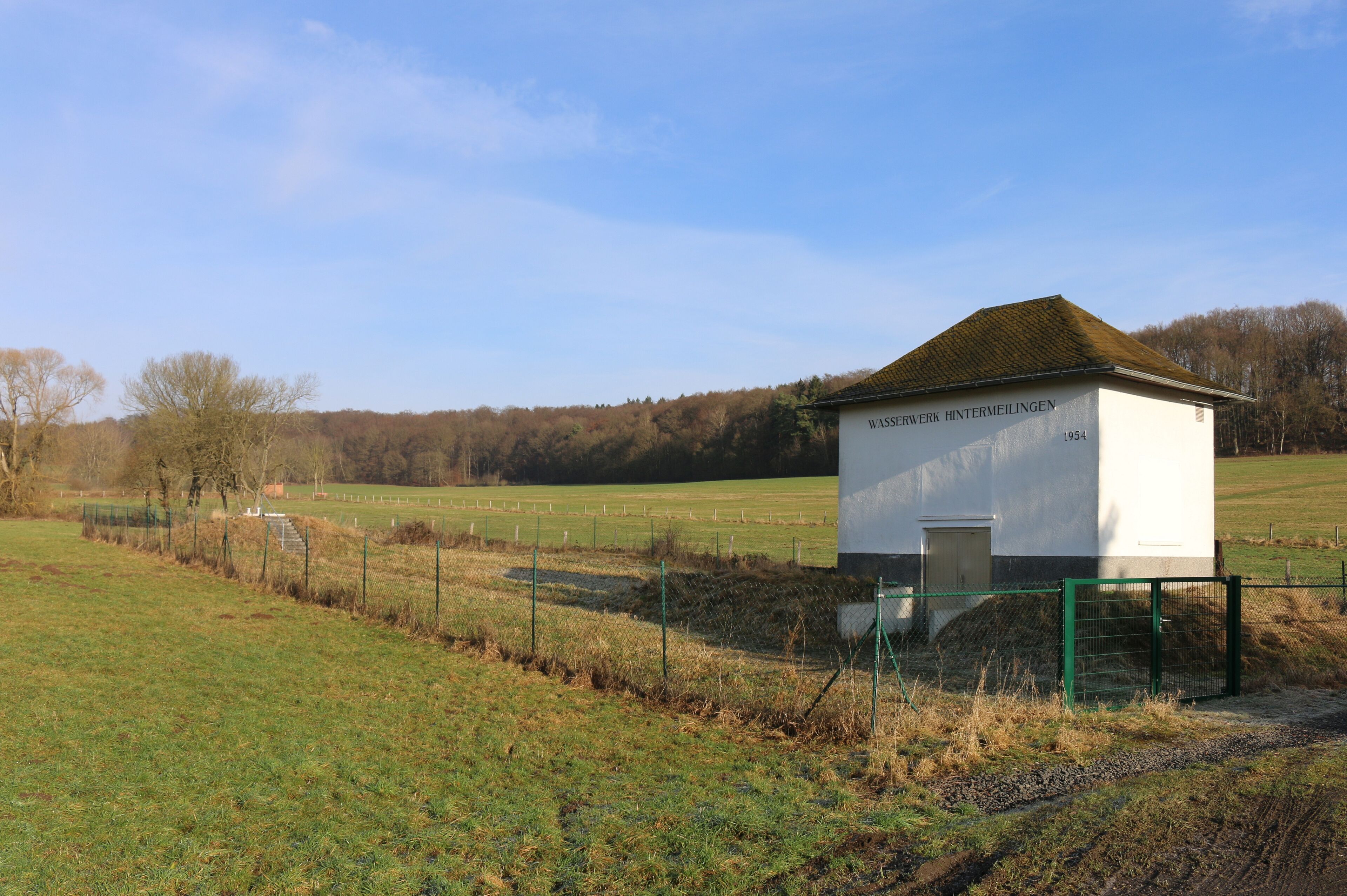 A small white building with a green roof labeled "WASSERWERK HINTERMEILINGEN 1954" in a rural field with a fence and trees in the background.