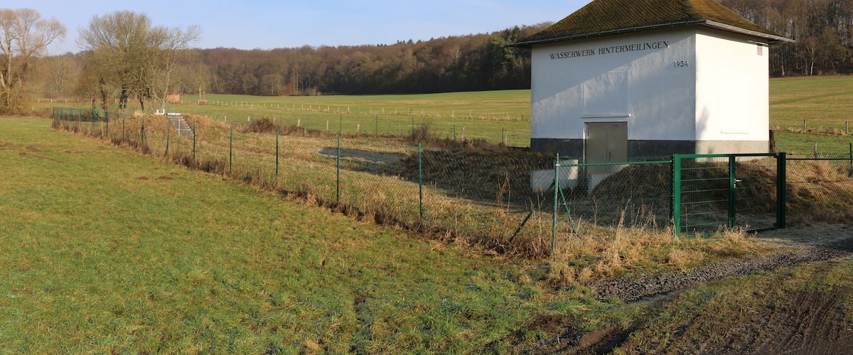 A small white building with a green roof labeled "WASSERWERK HINTERMEILINGEN 1954" in a rural field with a fence and trees in the background.