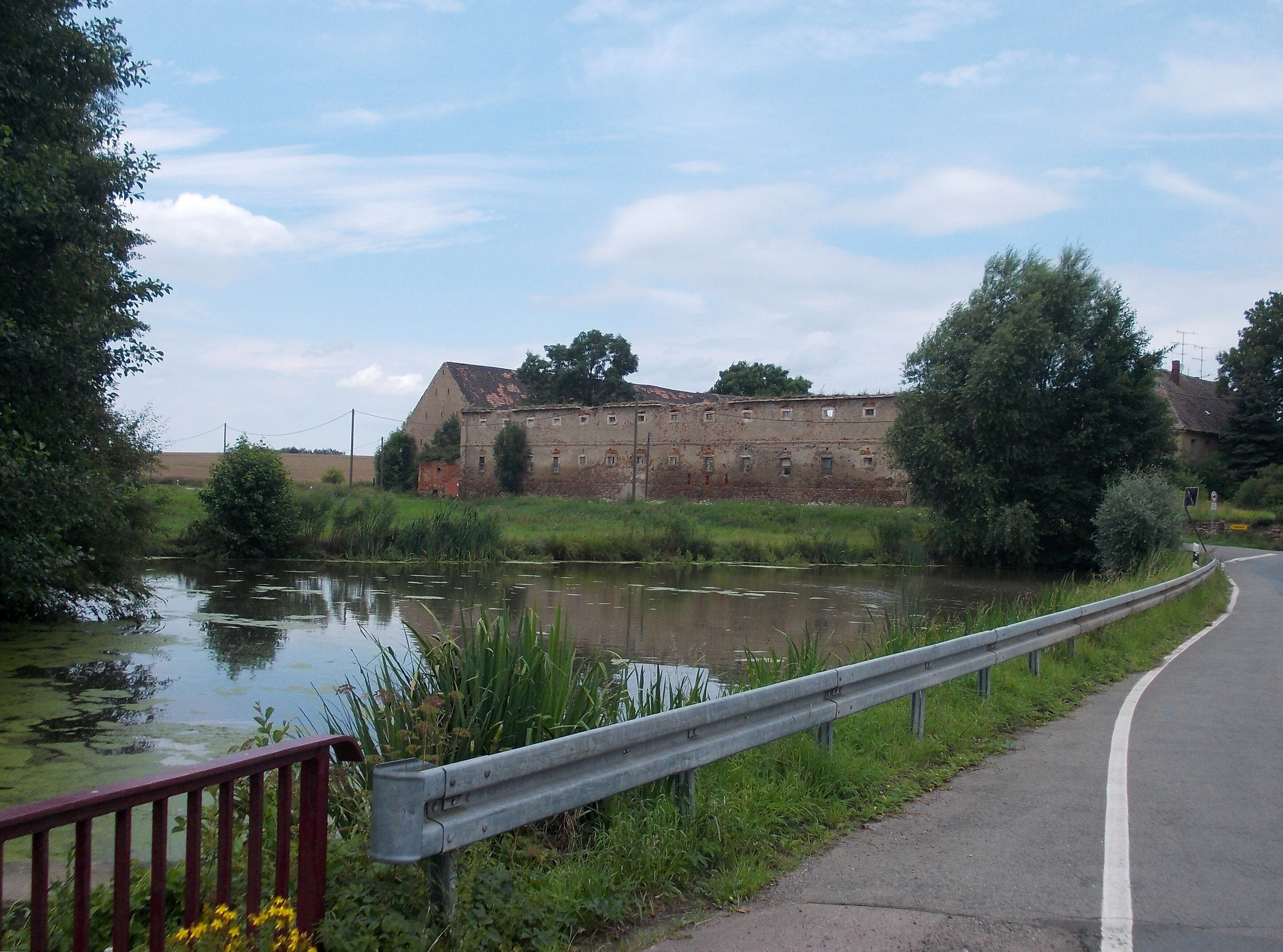 A rural landscape with a river, a dilapidated building, and a guardrail alongside a road.
