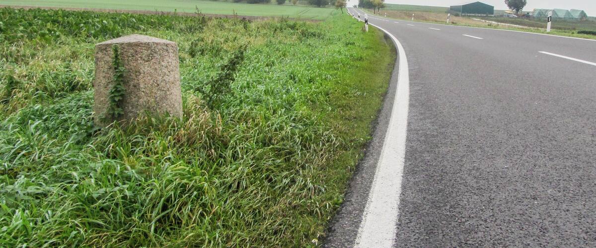 A rural road curving through a green field with a stone marker and distant trees.