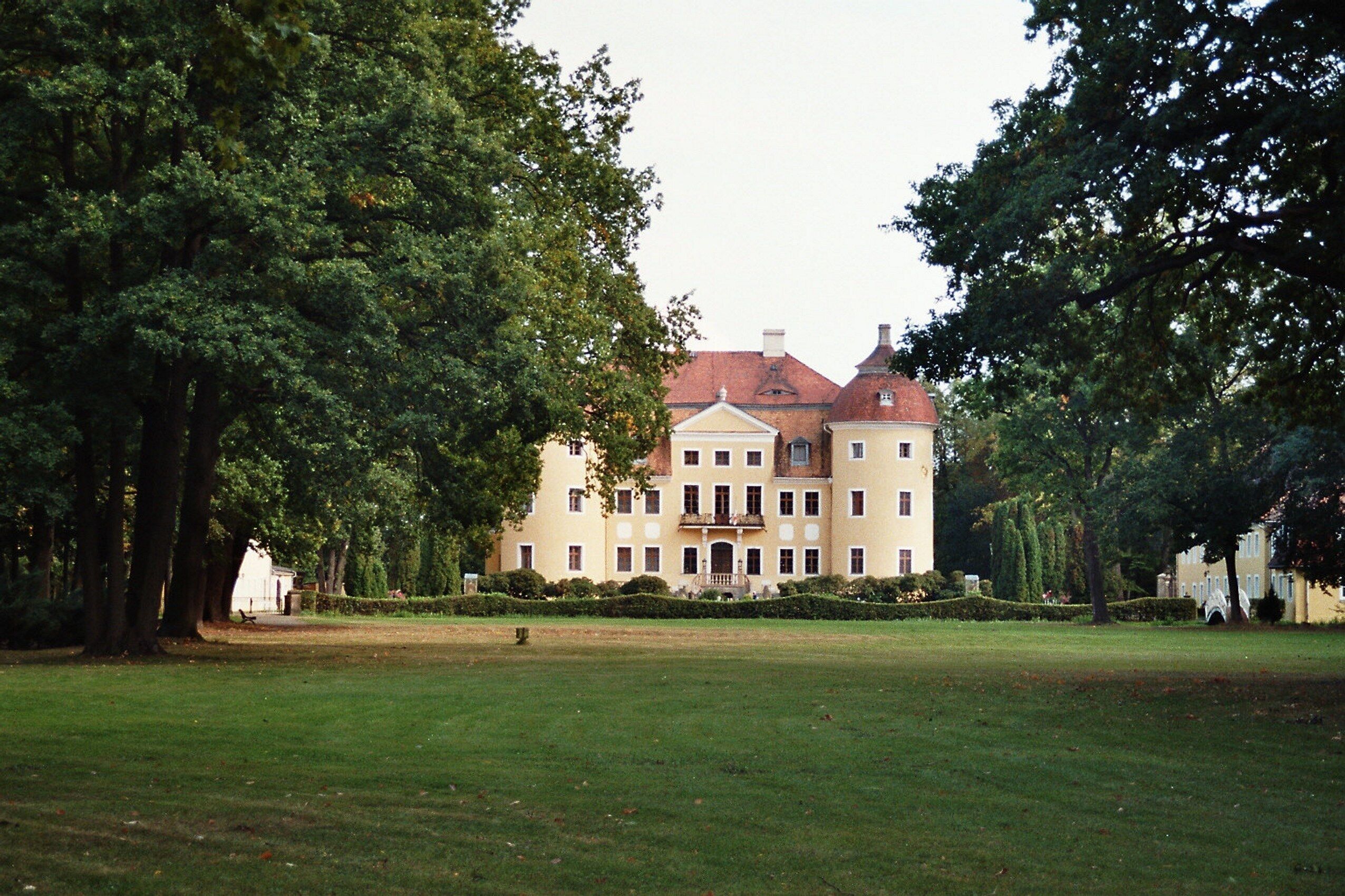 A large, historic building with a red roof, surrounded by lush green trees and a well-maintained lawn.
