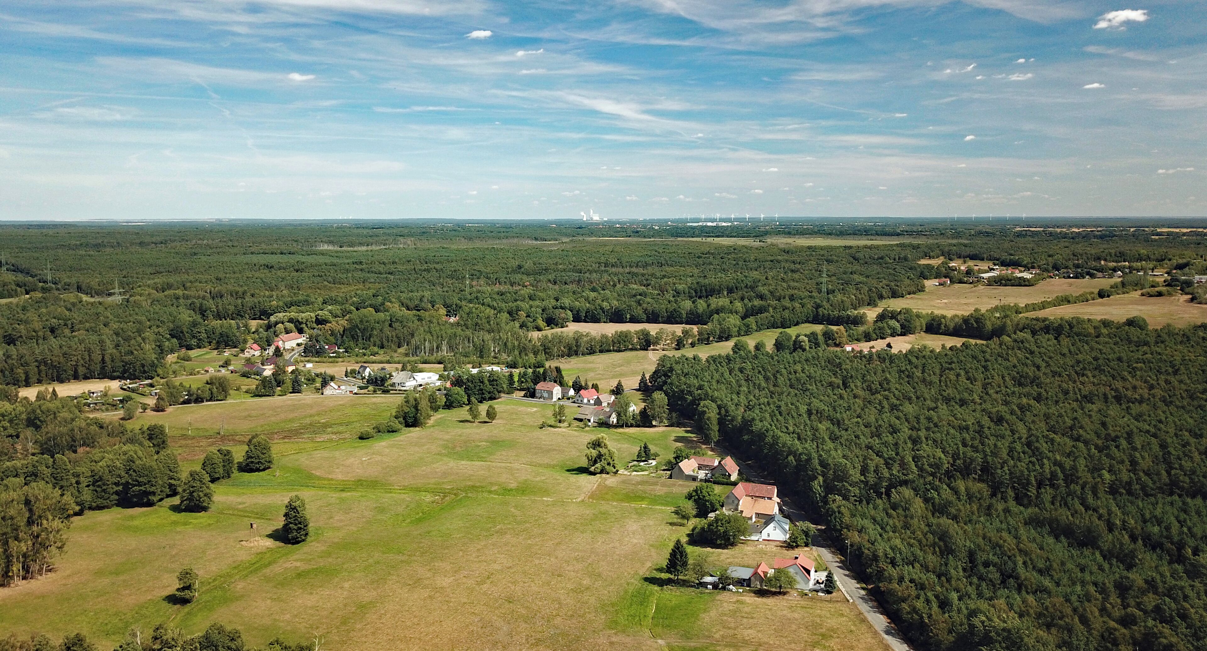 An aerial view of a rural landscape with scattered houses, dense forests, and distant wind turbines.