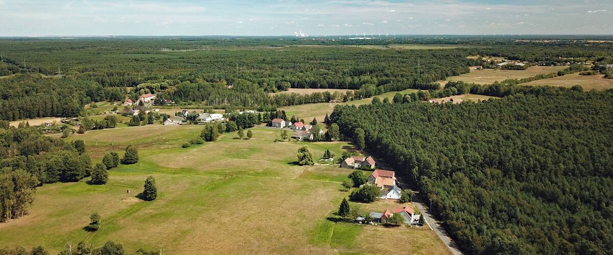An aerial view of a rural landscape with scattered houses, dense forests, and distant wind turbines.
