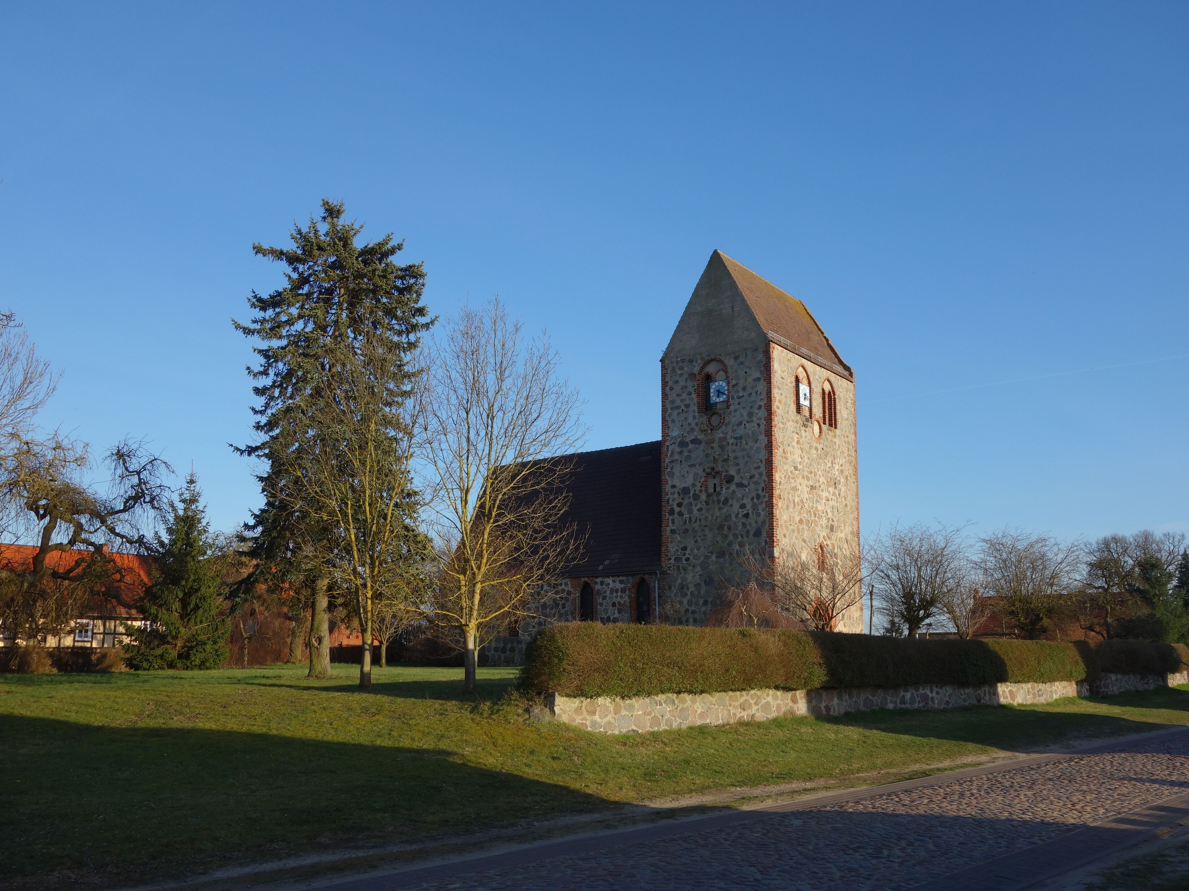 A stone church with a clock tower, surrounded by trees and a cobblestone path.