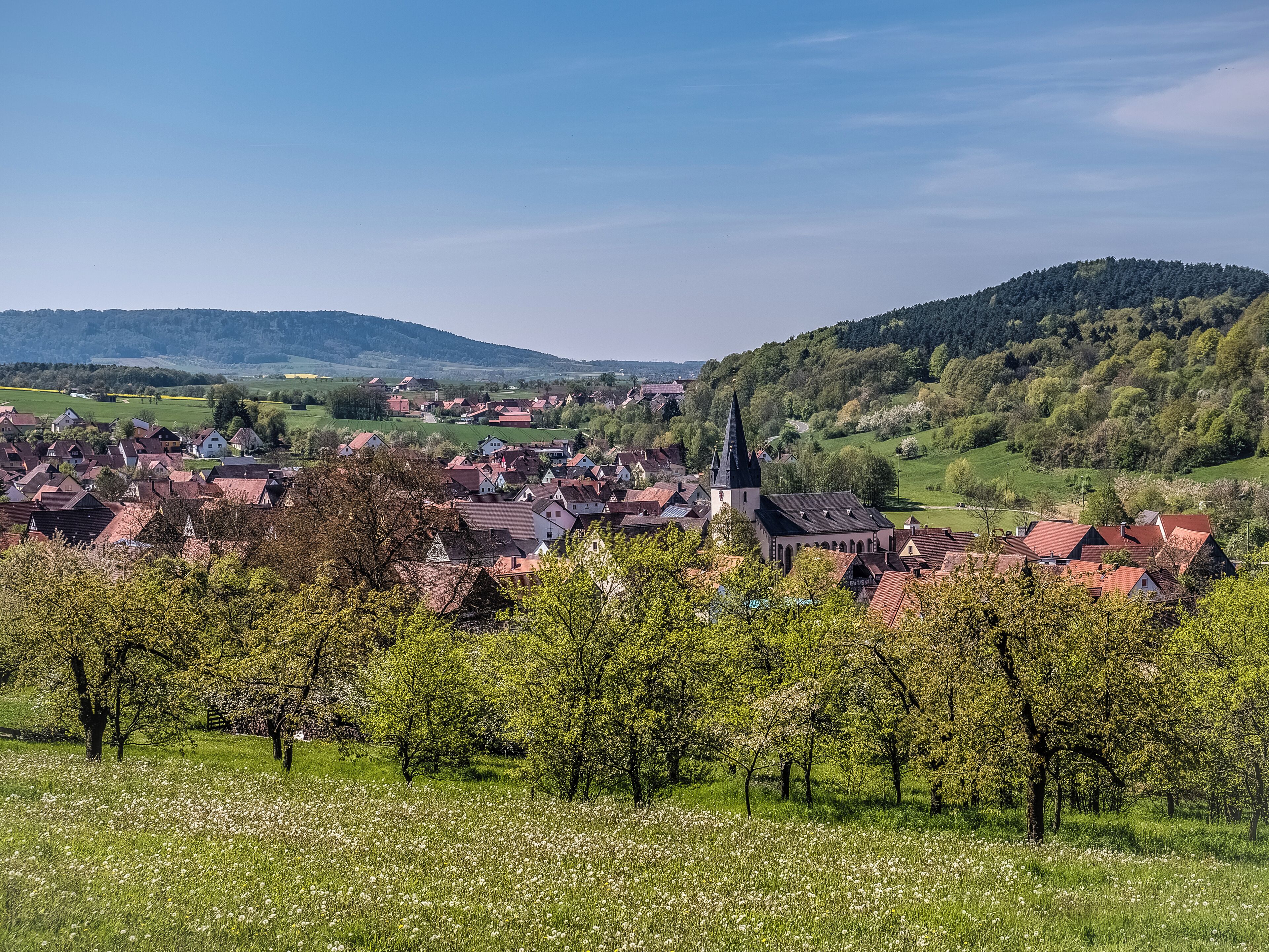 A village with a prominent church, surrounded by lush green hills and blooming trees.