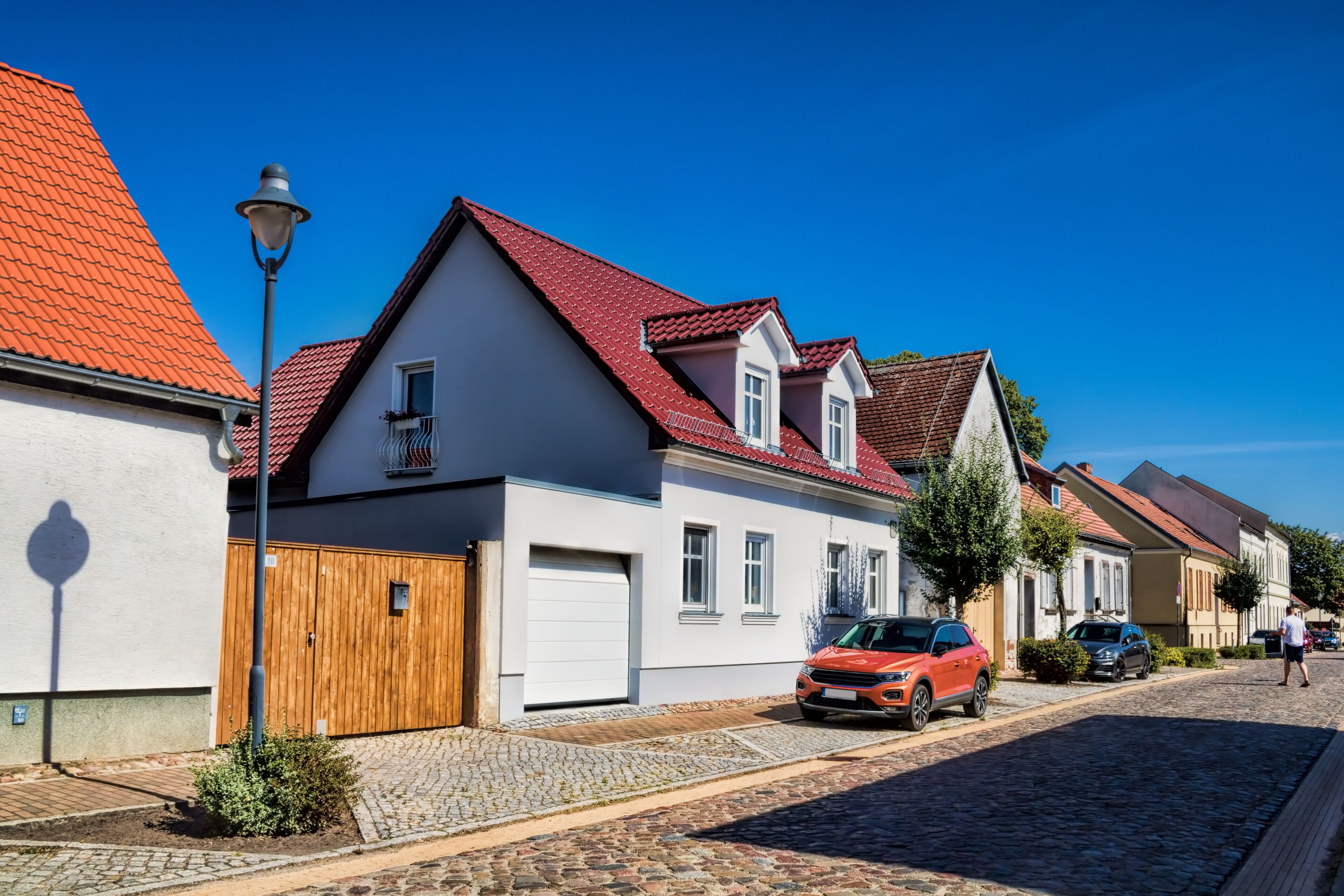 A cobblestone street lined with traditional houses, a wooden gate, and parked cars.