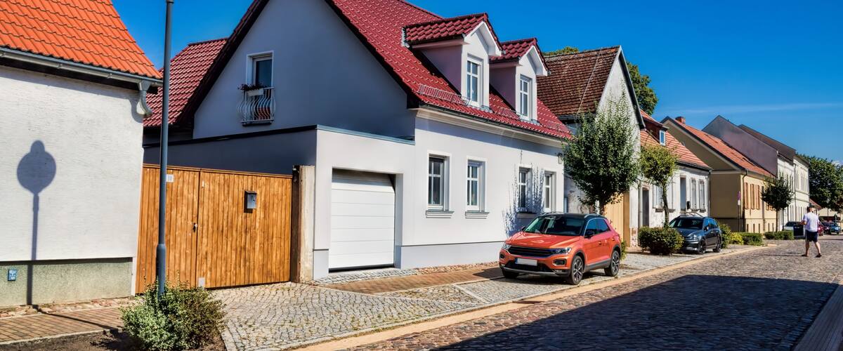 A cobblestone street lined with traditional houses, a wooden gate, and parked cars.