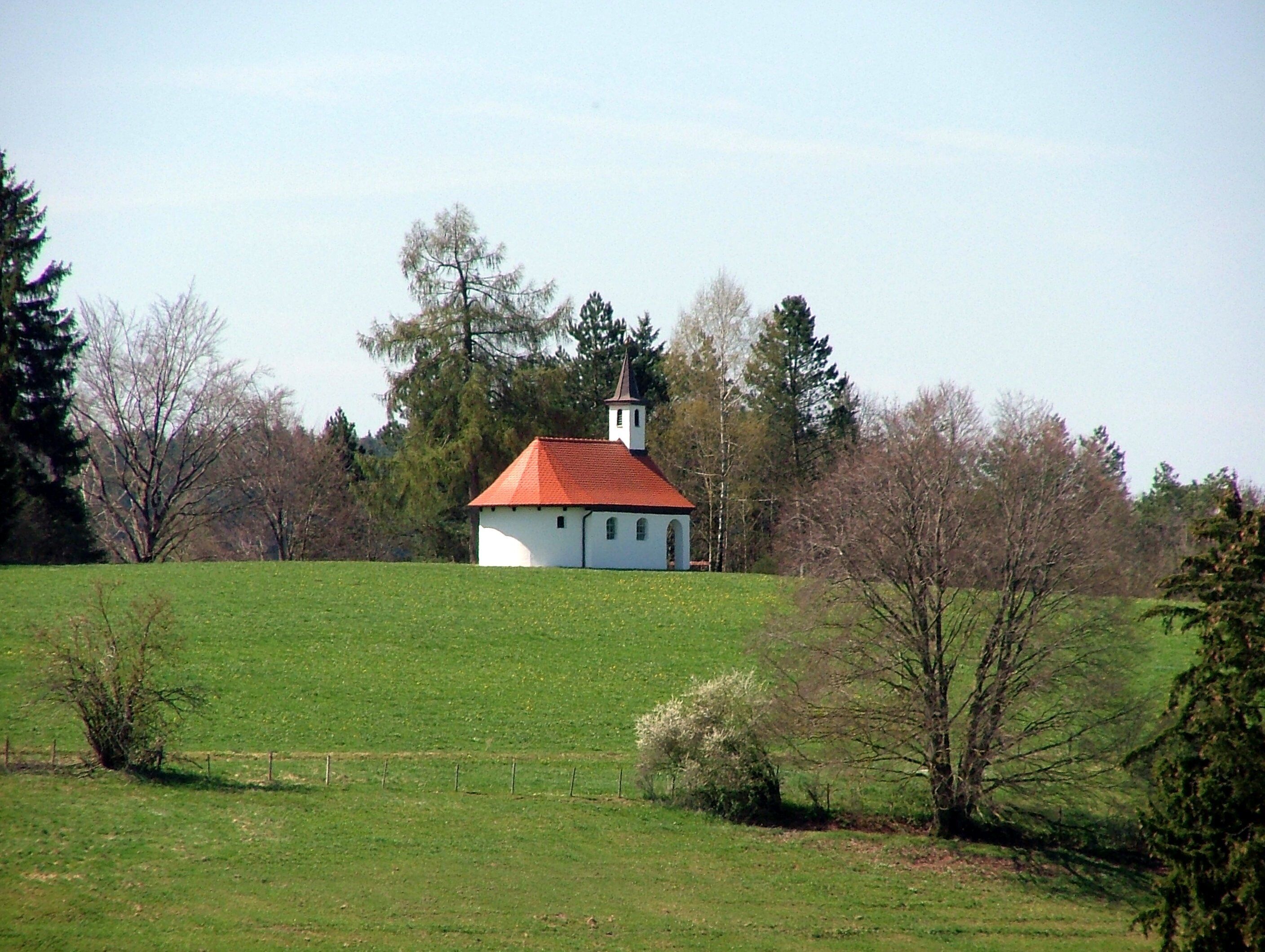 A white church with a red roof in a green field surrounded by trees.