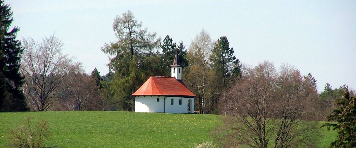 A white church with a red roof in a green field surrounded by trees.