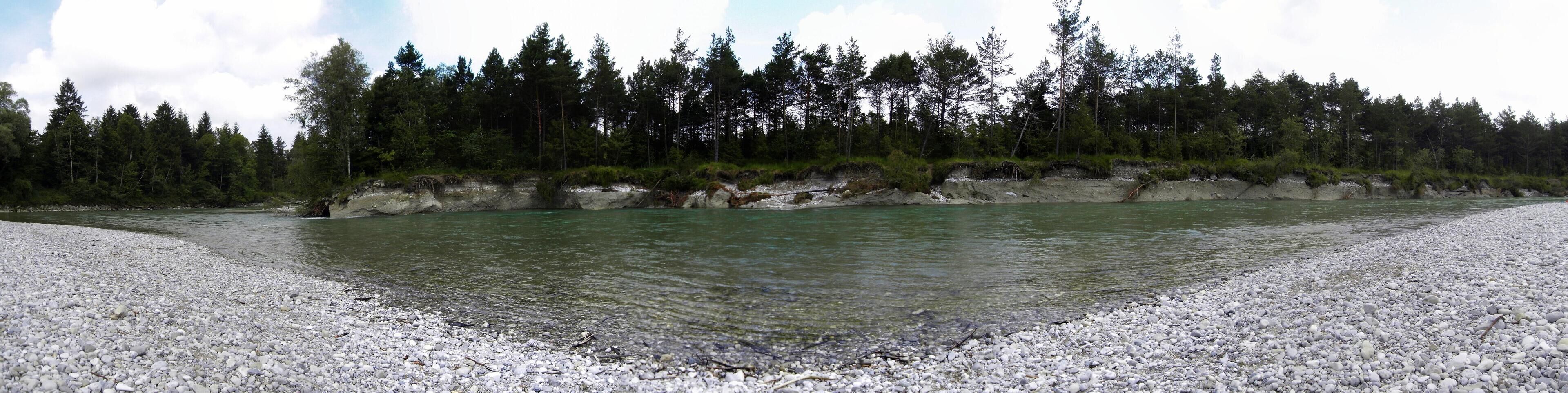 A river with clear water, surrounded by a rocky shoreline and dense forest.